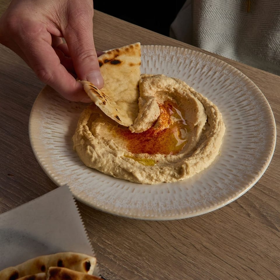 A hand dips a piece of pita bread into a plate of hummus drizzled with olive oil and paprika.