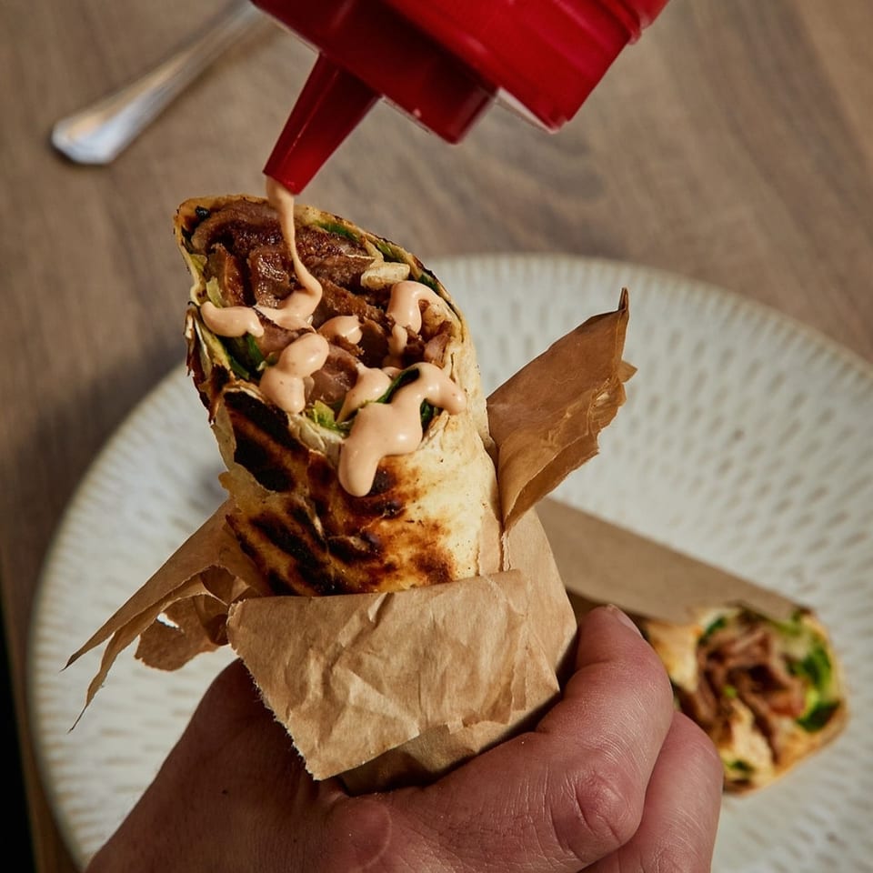 A hand holds a grilled wrap, drizzling it with sauce from a red squeeze bottle.
